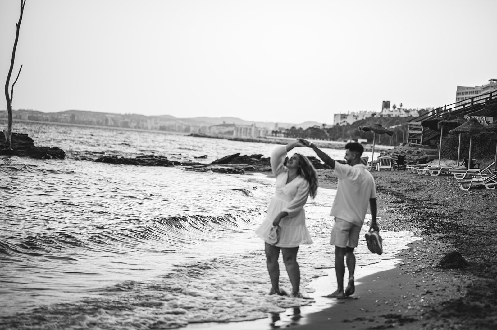 Dos personas caminan descalzas por una playa rocosa; una de ellas lleva un paraguas. La imagen está en blanco y negro.