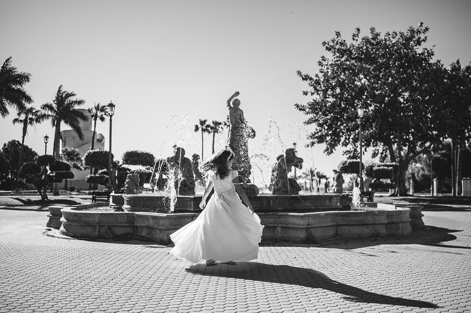 Una persona con un vestido blanco de pie en un parque soleado, cerca de una fuente, con árboles y palmeras al fondo
