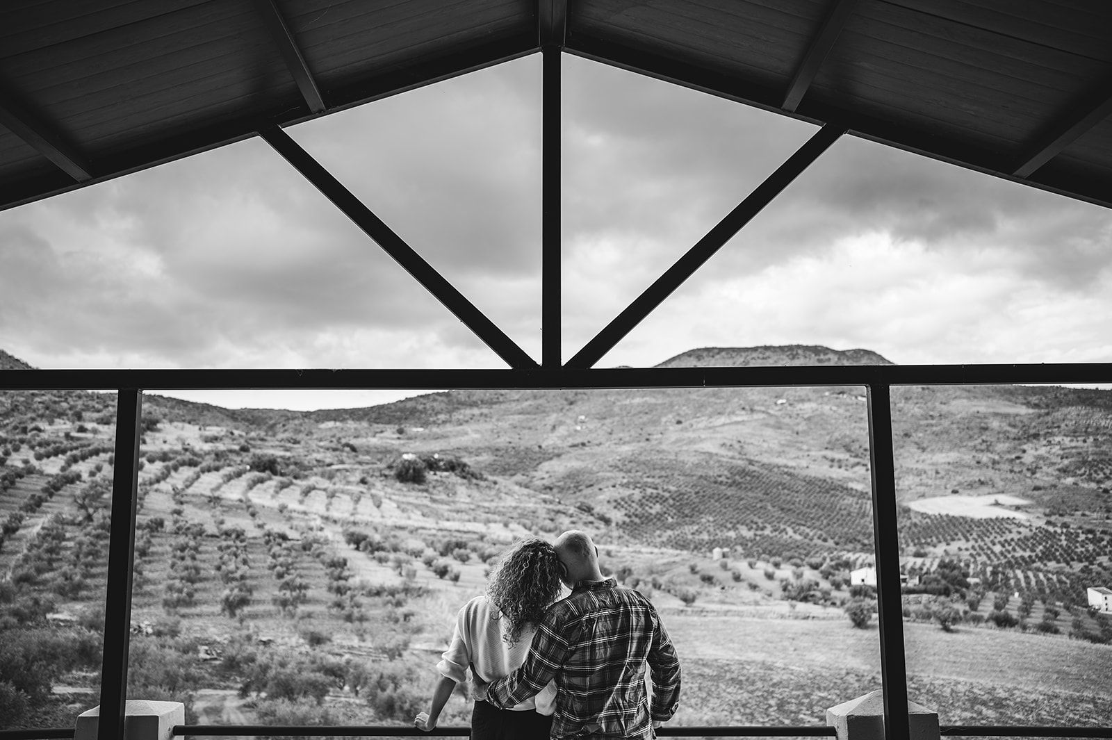 Dos personas sentadas bajo un techo, contemplando un paisaje montañoso en blanco y negro a través de grandes ventanales