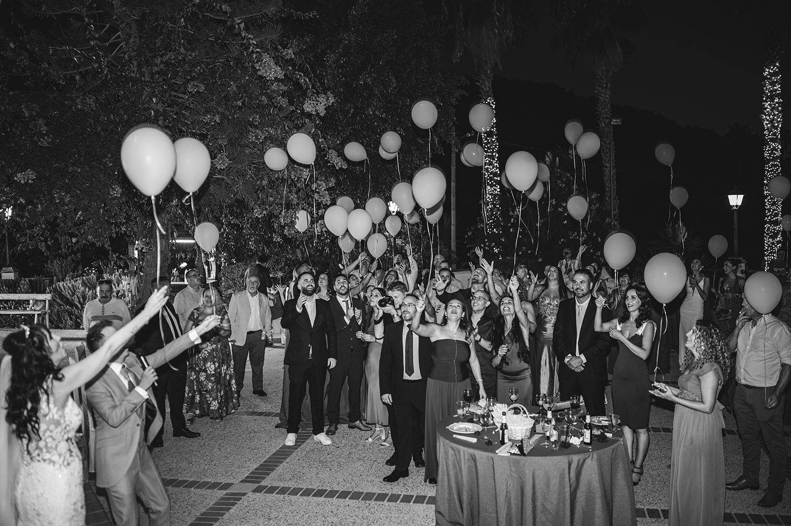 Una multitud en una celebración nocturna al aire libre soltando globos, con una mesa llena de flores delante
