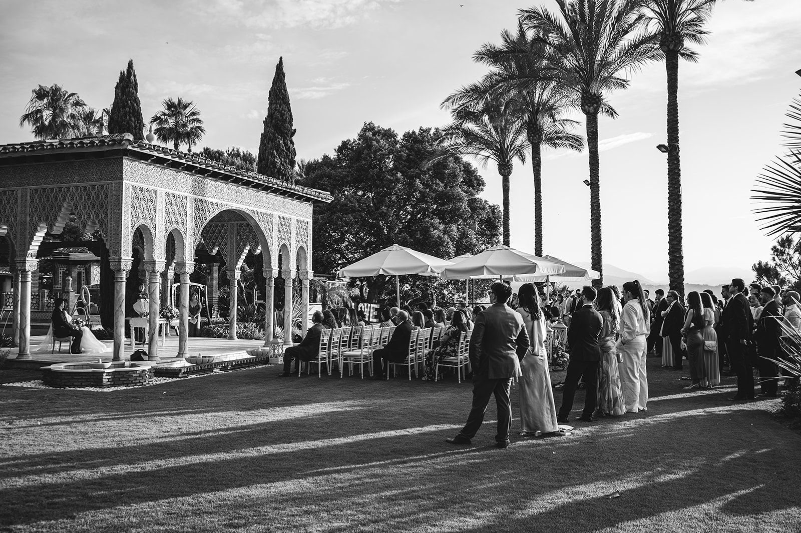 Recepción de boda al aire libre, con invitados charlando cerca de una villa, sombrillas y altas palmeras.