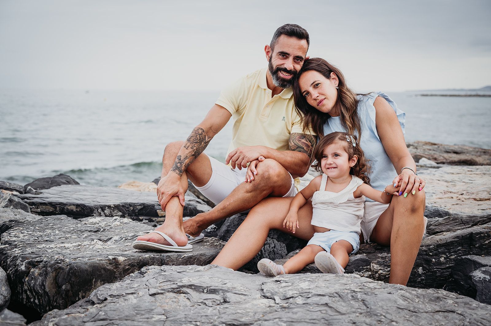 Una familia sentada en unas rocas junto al mar, sonriendo a la cámara con las olas de fondo