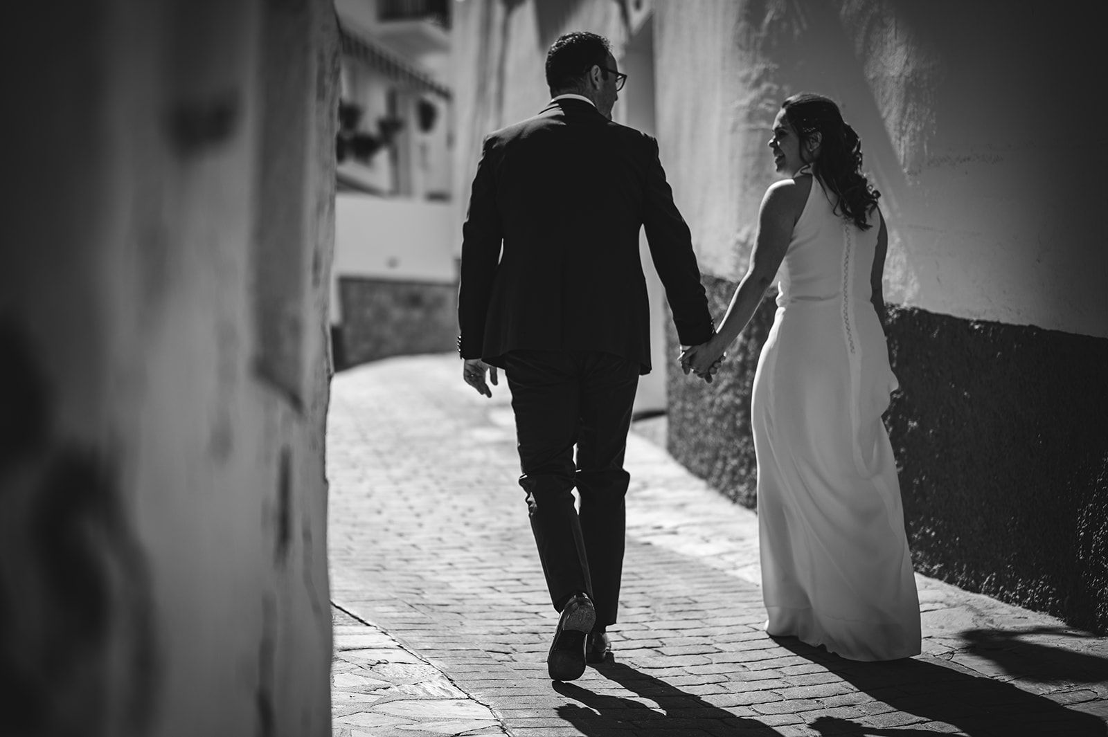 Una pareja vestida de gala camina de la mano por un callejón de piedra bañado por el sol.