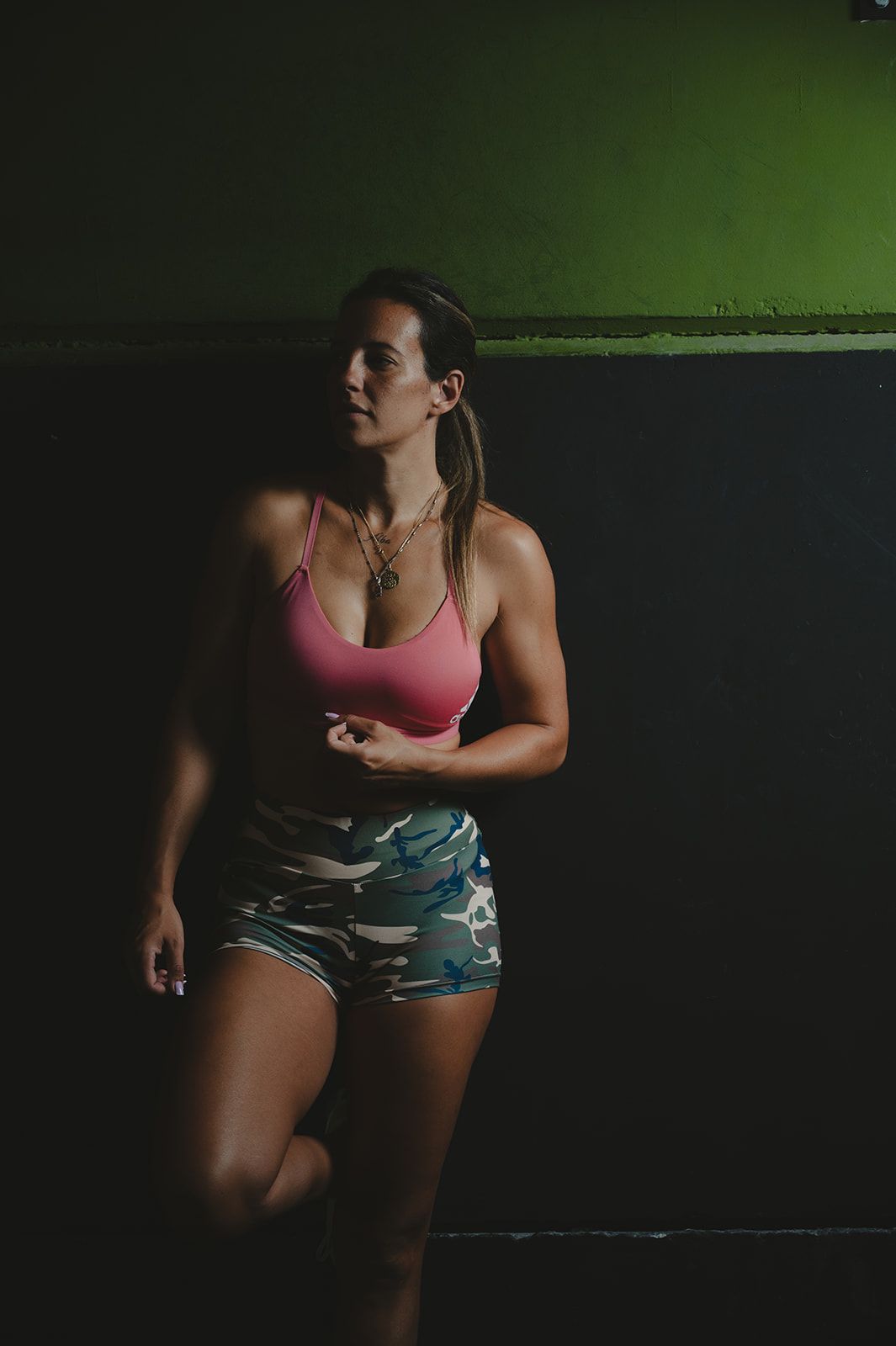 Mujer apoyada contra una pared, con un top rosa y pantalones cortos estampados bajo una luz verde
