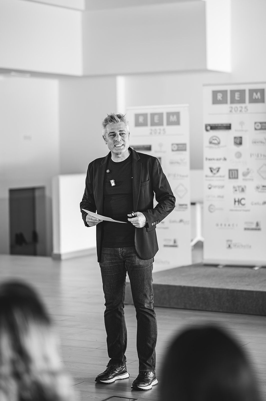 Un ponente sobre el escenario, con una chaqueta negra, dirigiéndose al público en un evento en un espacio cerrado.