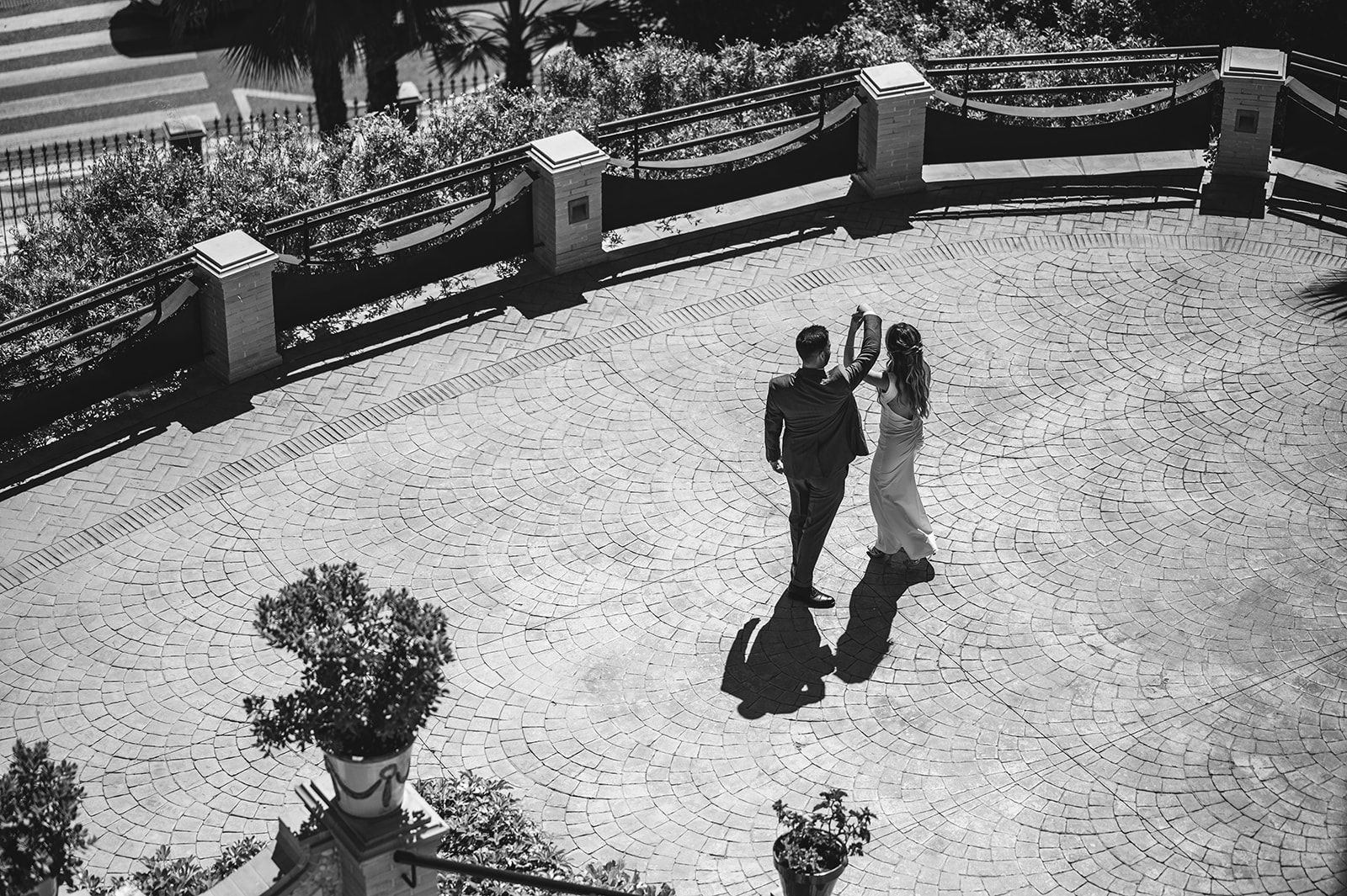 Dos personas de pie, muy juntas, en un patio soleado con sombras alargadas y una barandilla de piedra curva.