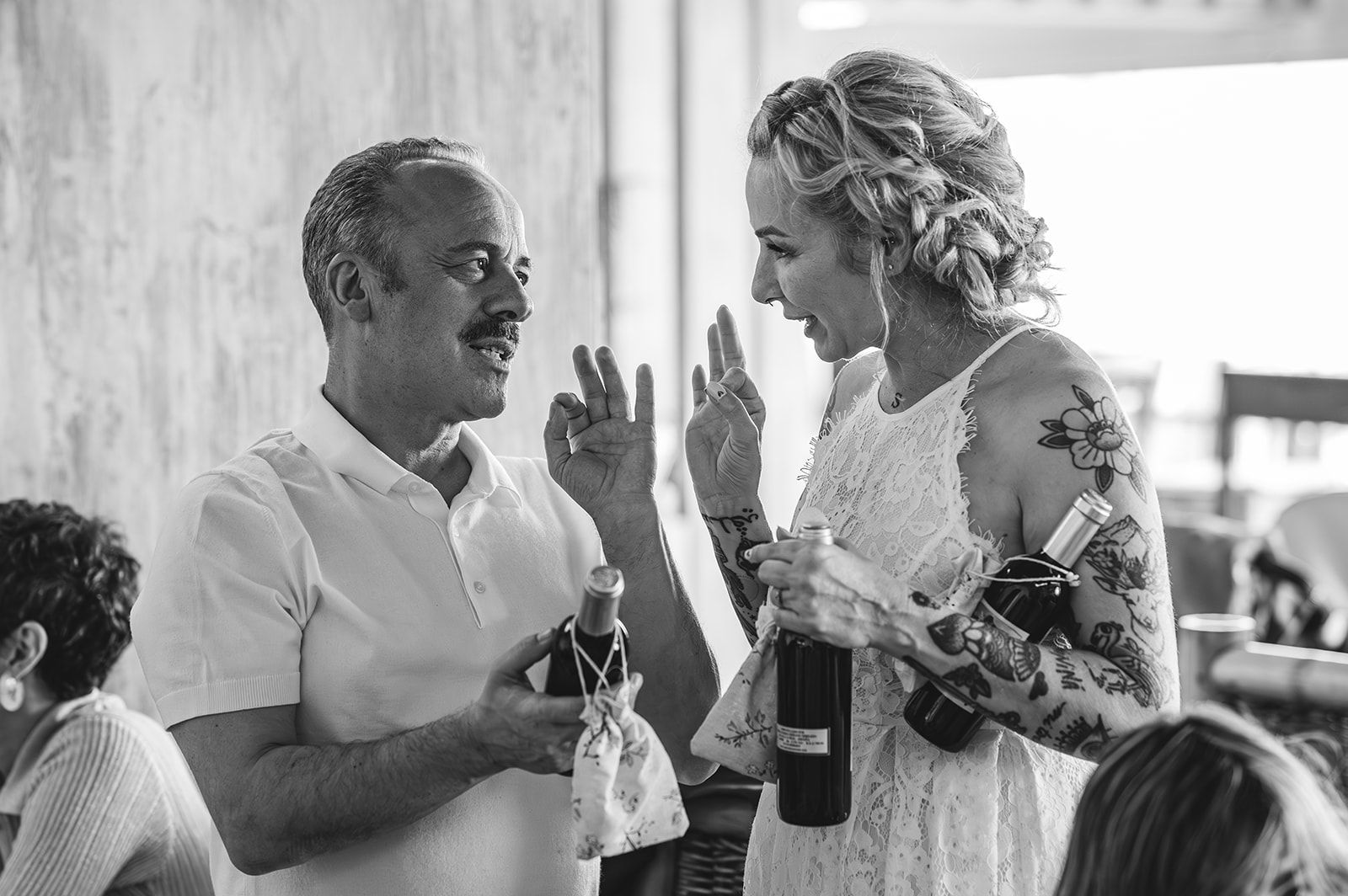 Dos personas sonríen y conversan en una reunión social, con bebidas en la mano y haciendo gestos con los dedos levantados