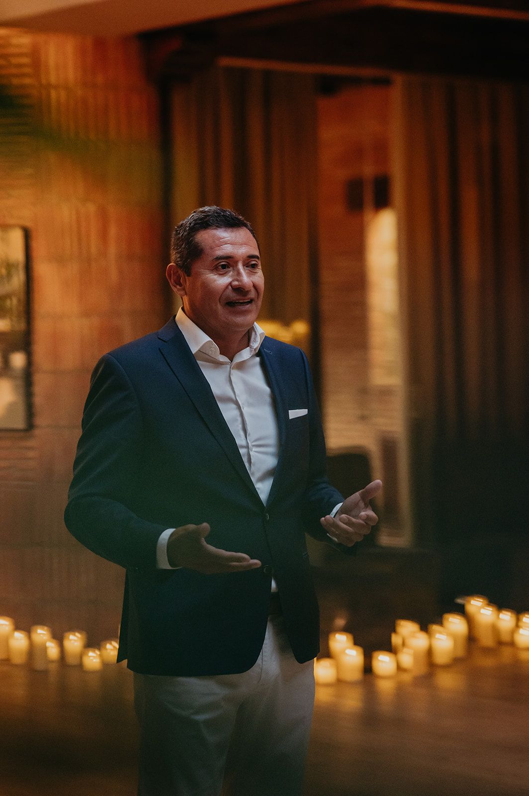 Hombre hablando en un escenario, con una chaqueta oscura, un fondo iluminado por velas cálidas y una expresión concentrada
