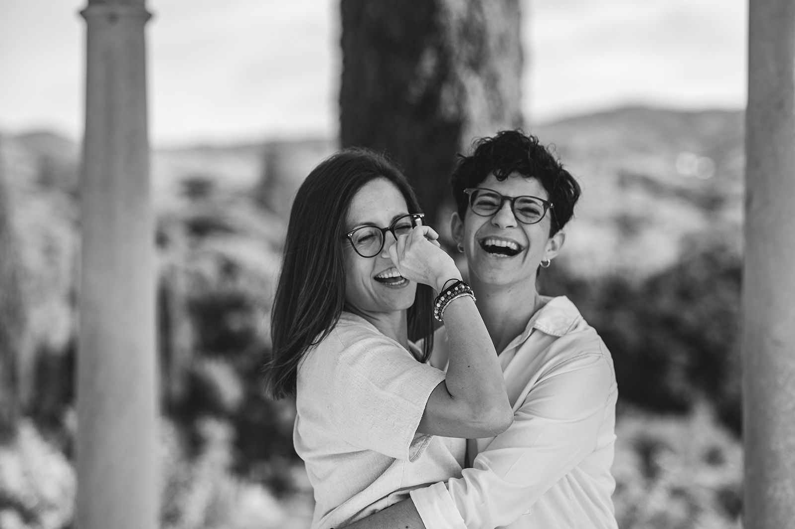 Dos personas riéndose y abrazándose al aire libre bajo una estructura con sombra, en blanco y negro.