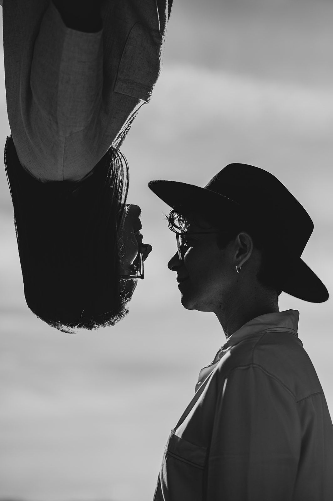 Dos personas de perfil frente a frente, una con sombrero, sobre un fondo de cielo nublado y agua.