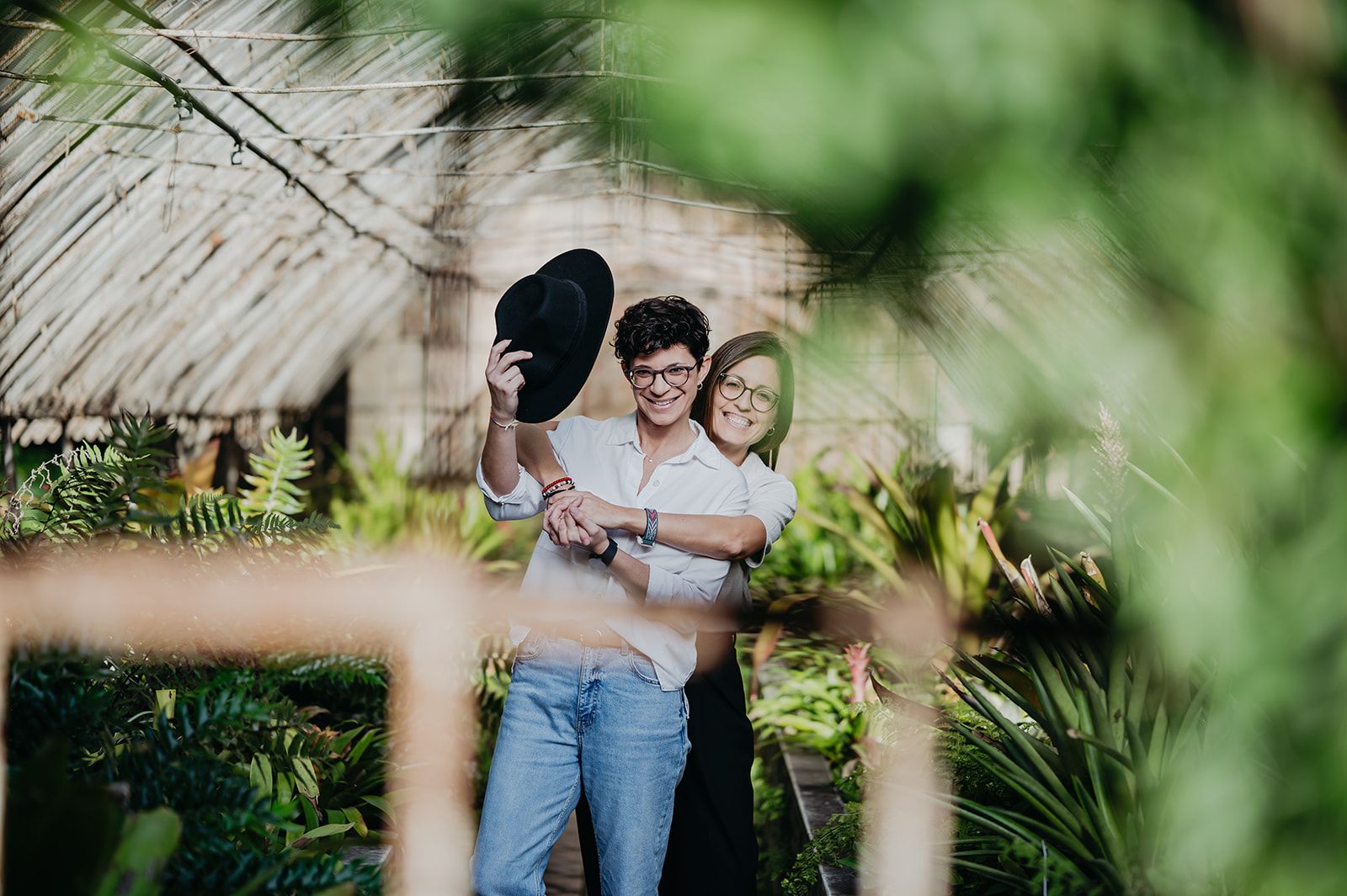 Una pareja sonriente sostiene un sombrero negro en un exuberante jardín, rodeada de vegetación verde