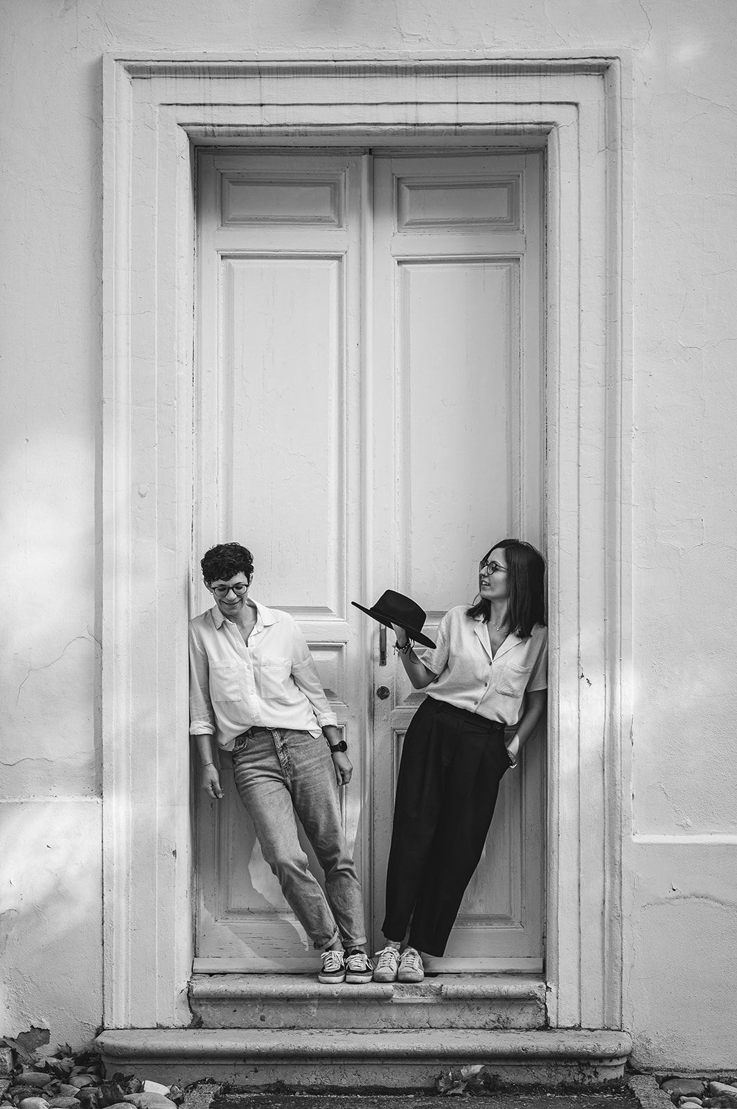 Dos personas apoyadas en el marco de una puerta; una lee un libro mientras la otra observa.