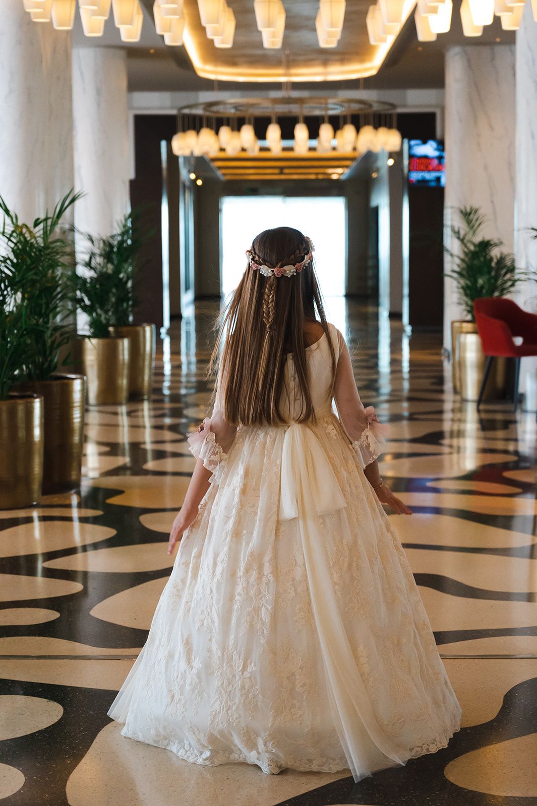 Una niña con un vestido blanco de encaje caminando por un majestuoso pasillo iluminado por lámparas de araña, con plantas en macetas y suelos estampados