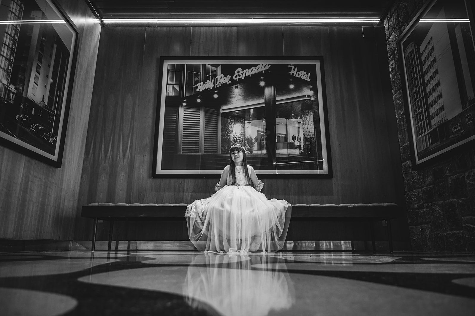 Una mujer con un vestido blanco, sentada sola en una zona de salón o escenario con poca iluminación.