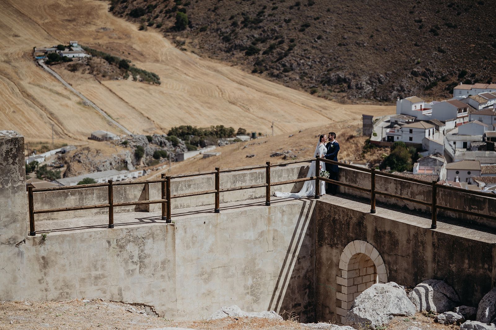 Vista desde un puente sobre un valle seco entre colinas, con algunas personas y edificios blancos a lo lejos