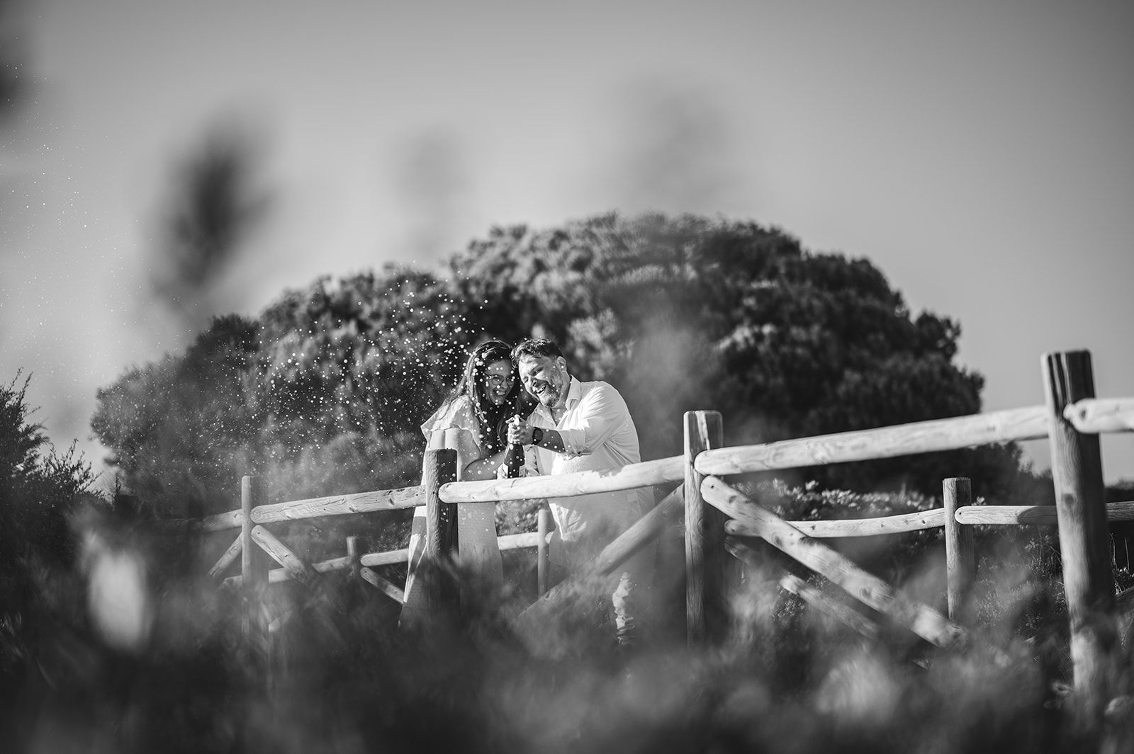 Pareja en blanco y negro abrazándose detrás de una valla de madera, en un entorno al aire libre con césped