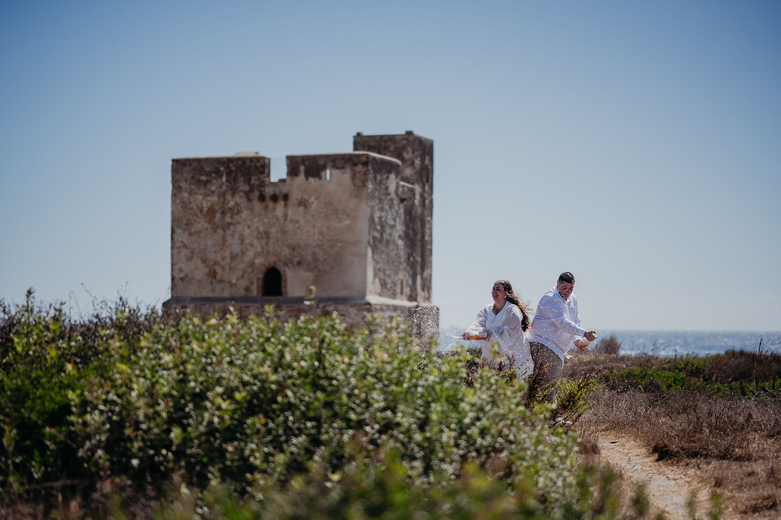 Dos personas paseando cerca de una antigua torre de piedra en un campo costero soleado
