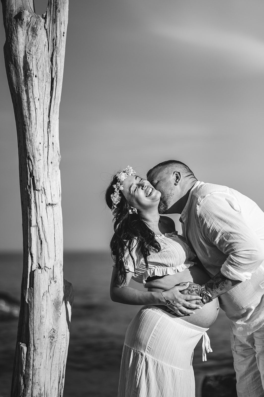 Una pareja abrazándose y besándose junto a un árbol en una playa, en blanco y negro.