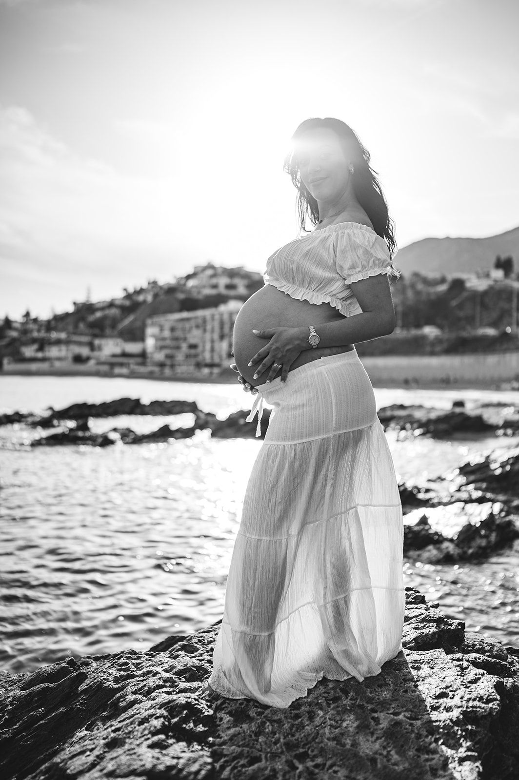 Mujer embarazada con un vestido blanco, de pie sobre unas rocas junto al mar al atardecer