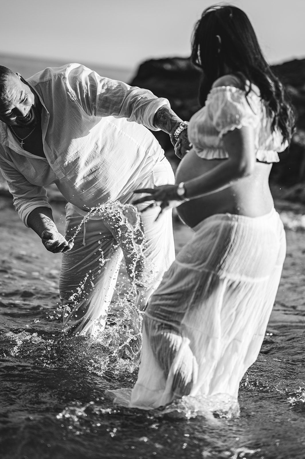 Dos personas chapoteando en aguas poco profundas; una, vestida de blanco, tiende la mano hacia la otra.