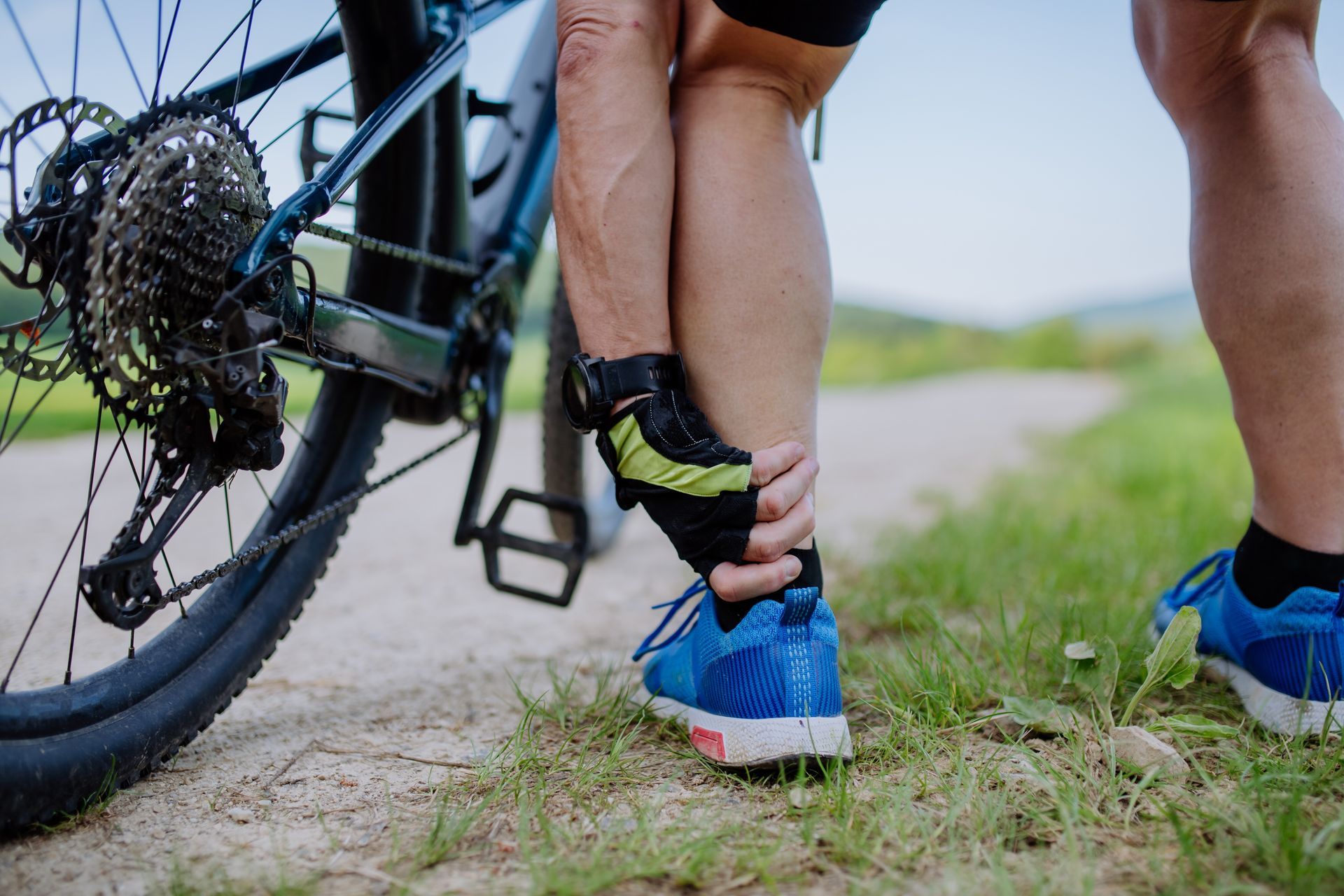 Cyclist holding their ankle on a dirt path beside their bicycle, showing possible injury.