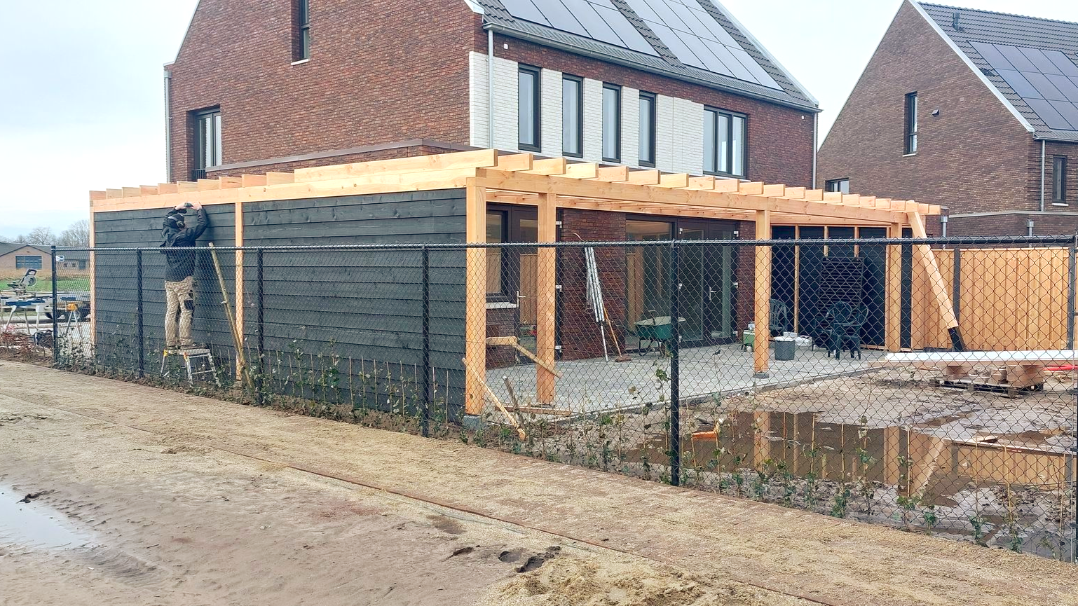Een houten pergola, bevestigd aan een bakstenen huis, naast een zwart hek.