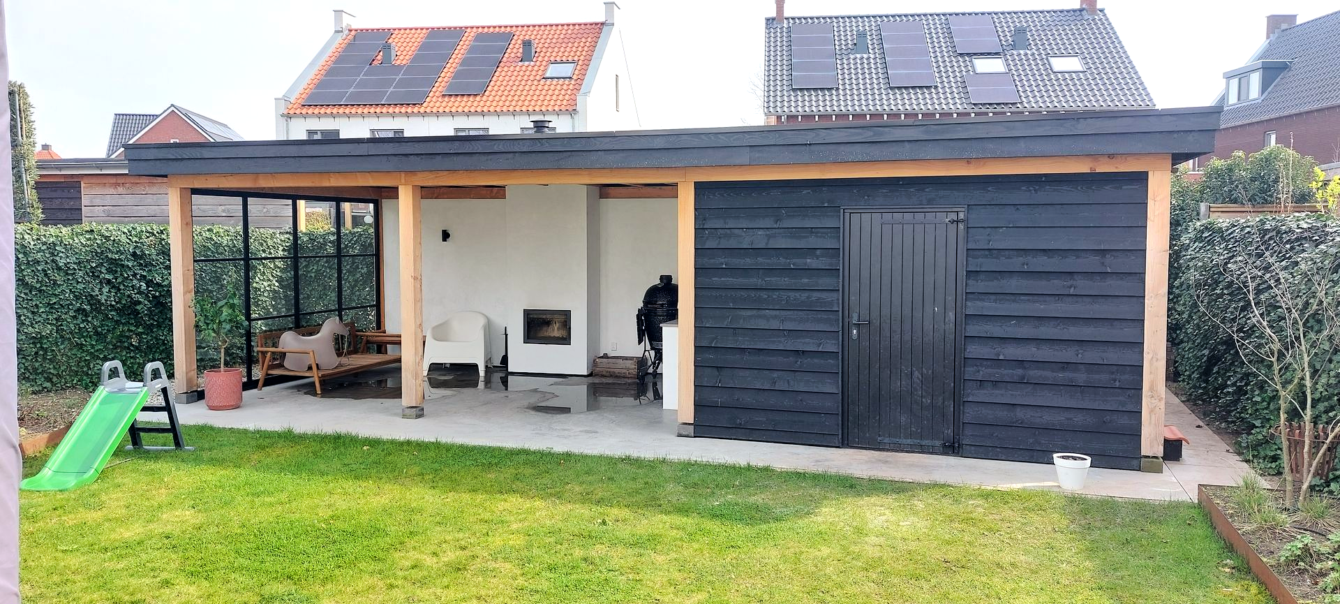 Terras in de achtertuin met een glazen zithoek, een open haard en een berging, met een groene glijbaan op het gras.