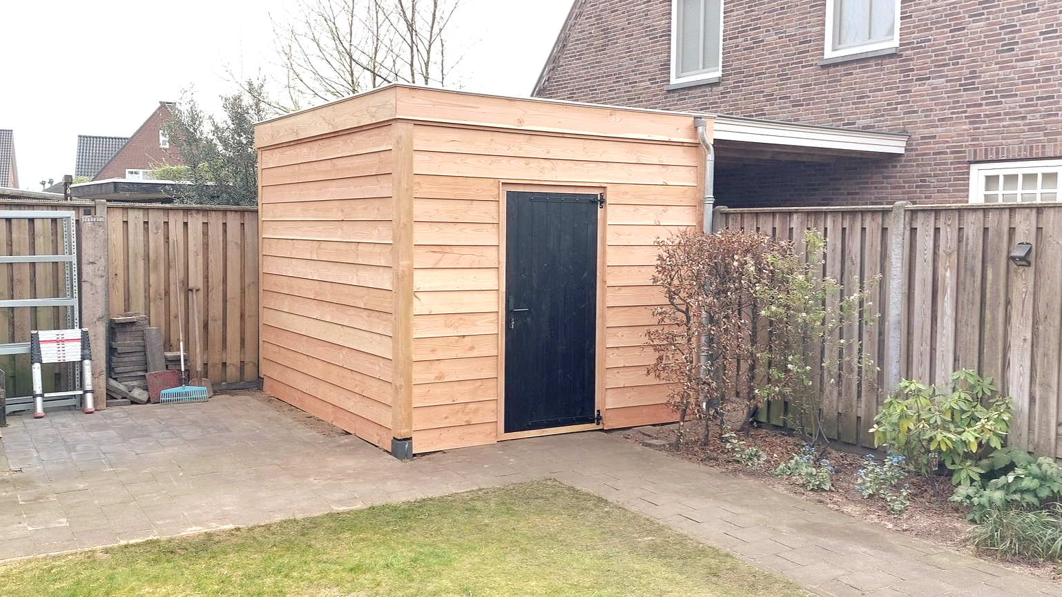 Houten schuur met zwarte deur in een achtertuin, naast een houten schutting en een huis.
