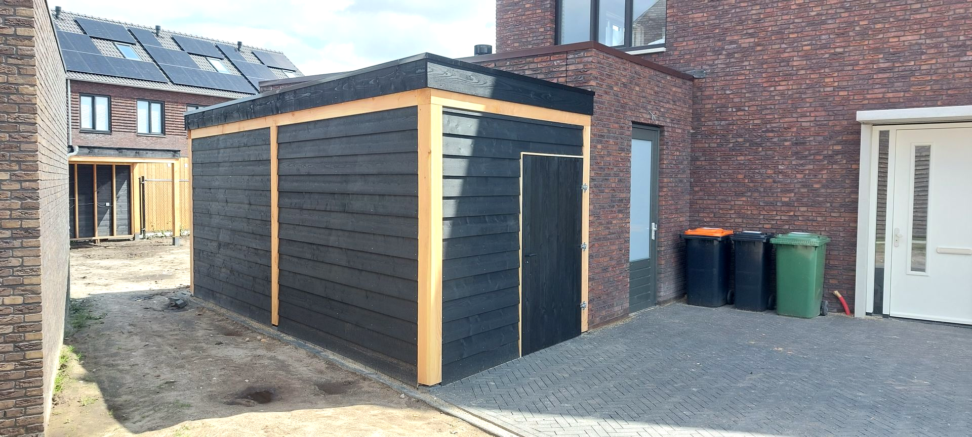 Zwarte schuur met lichte houten afwerking naast een bakstenen gebouw; drie afvalcontainers staan ​​er vlakbij.
