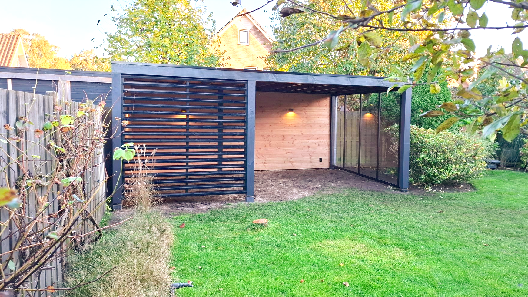Een pergola met zwarte houten frame in een achtertuin. Voorzien van horizontale latten aan één zijde en verlichting.