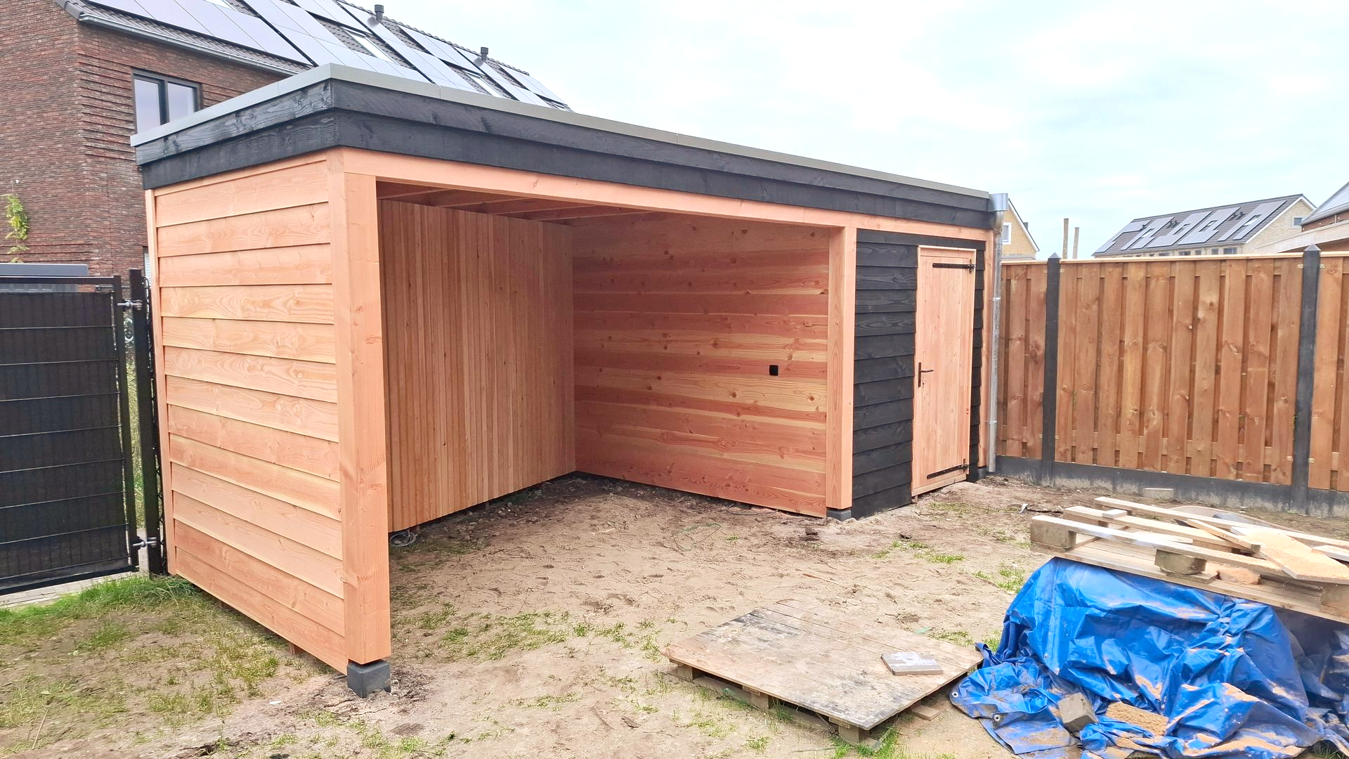 Houten carport met een open zijkant, een kleine deur en een donker dak. De carport staat in een achtertuin.
