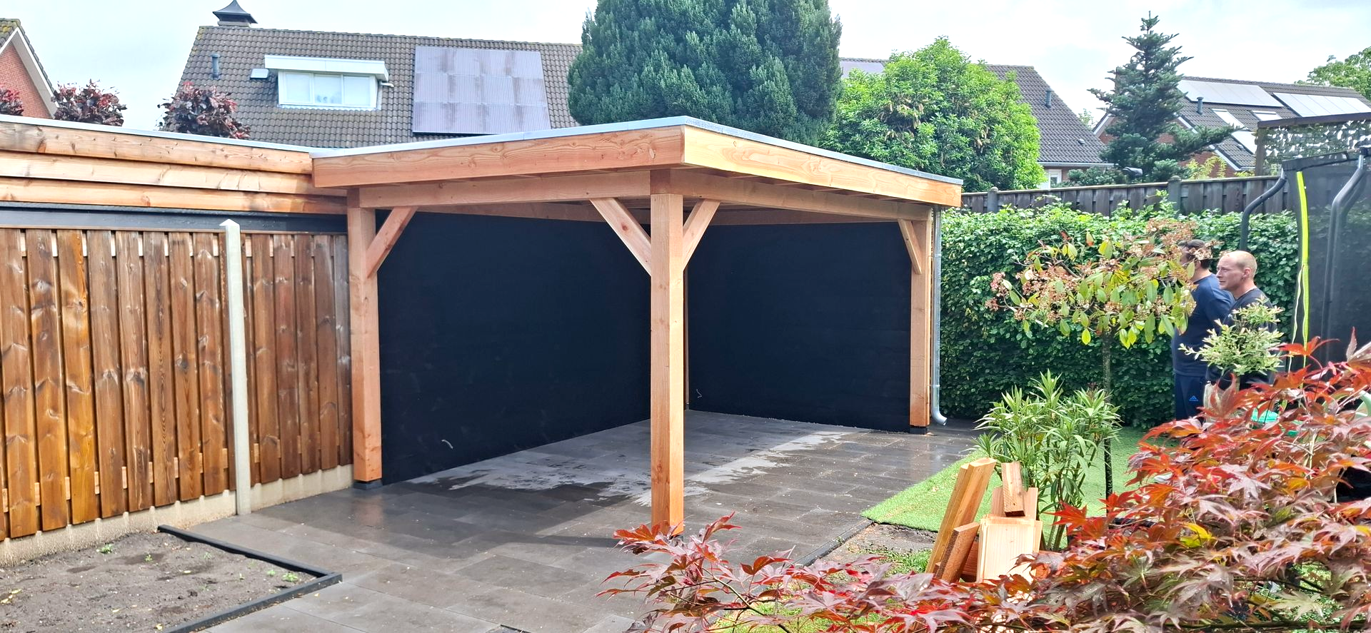 Houten carport naast een houten schutting met zwarte gevelbekleding. Op de achtergrond is iemand aan het werk.