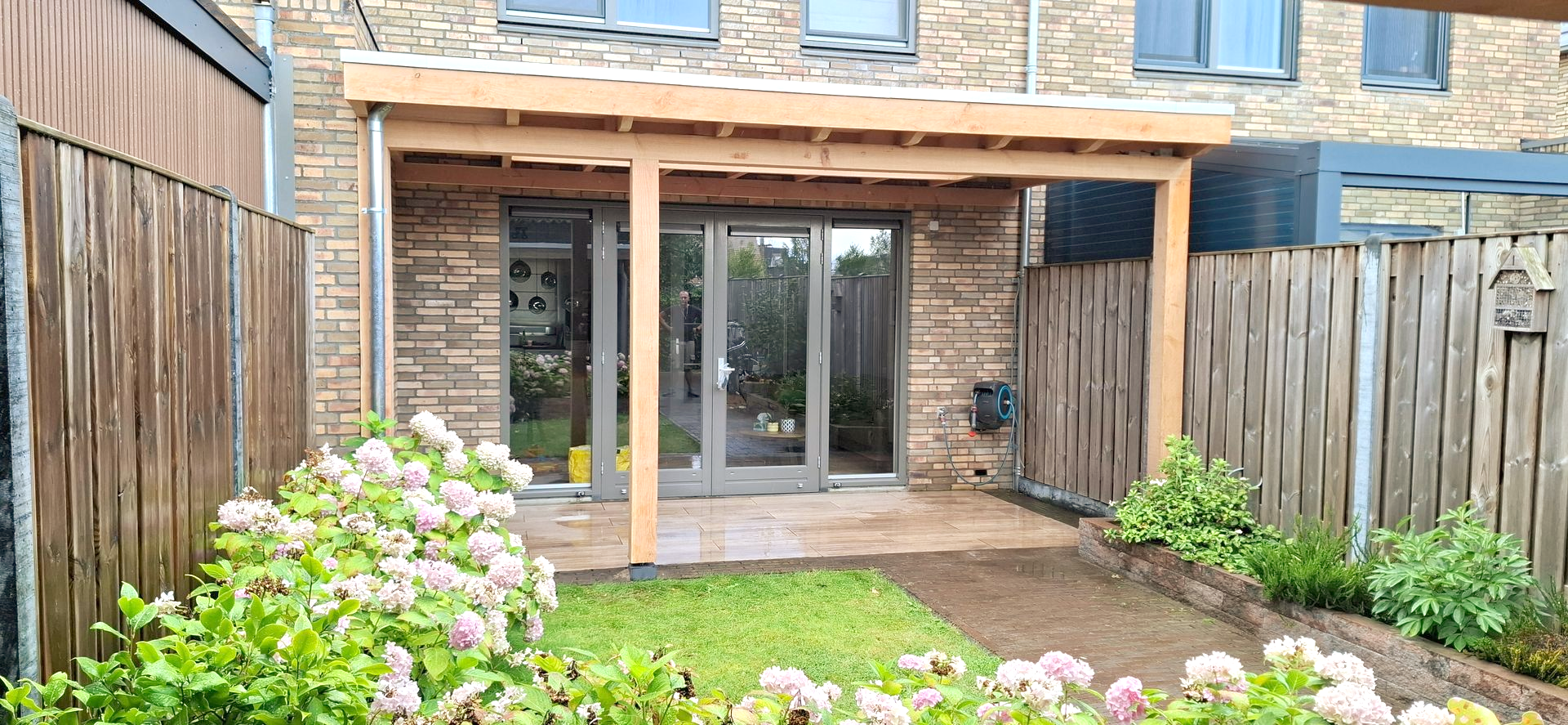 Terras in de achtertuin met houten pergola, bakstenen muur en groen gazon.