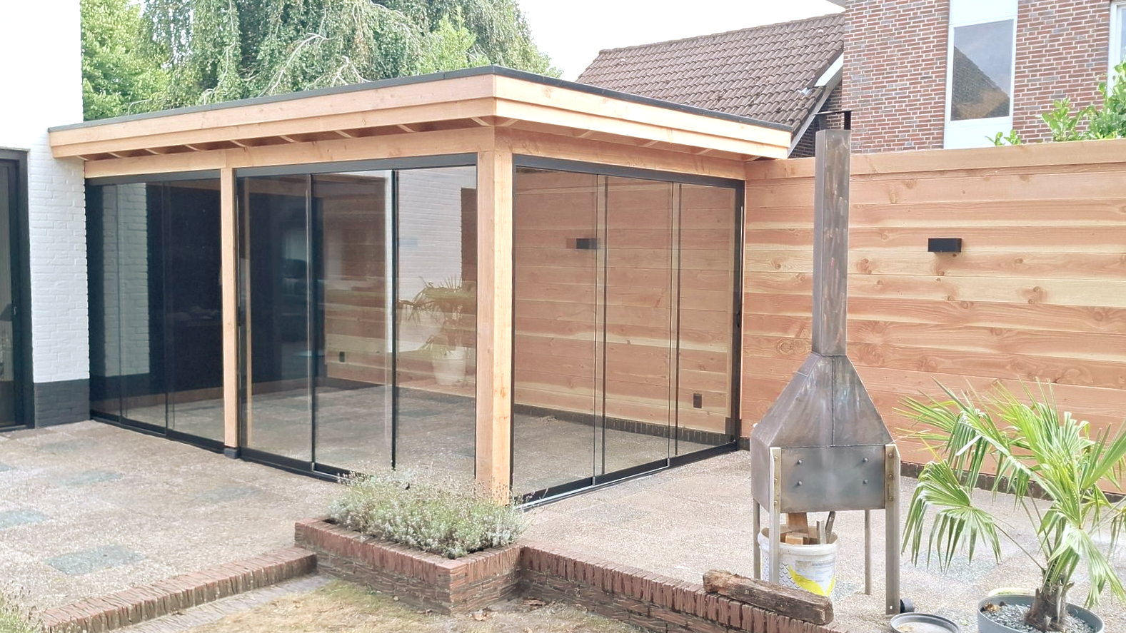 Houten terrasoverkapping met glazen wanden, grindterras, bakstenen rand en buitenhaard.