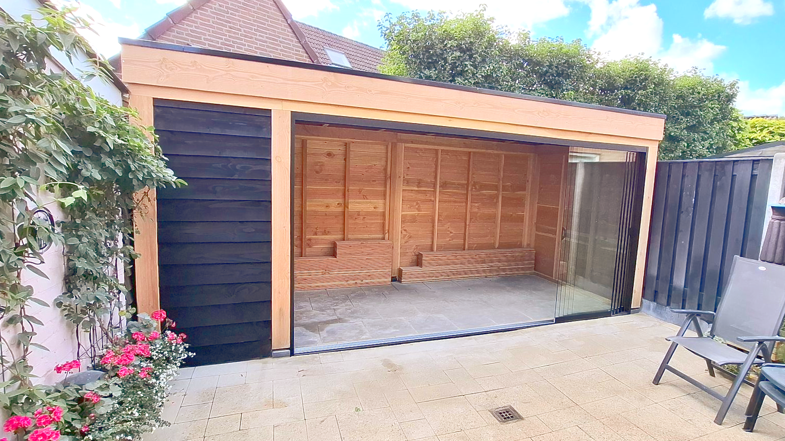 Open houten garage met grindvloer, gelegen in een achtertuin. Uitgevoerd in donkergrijs en naturel hout.