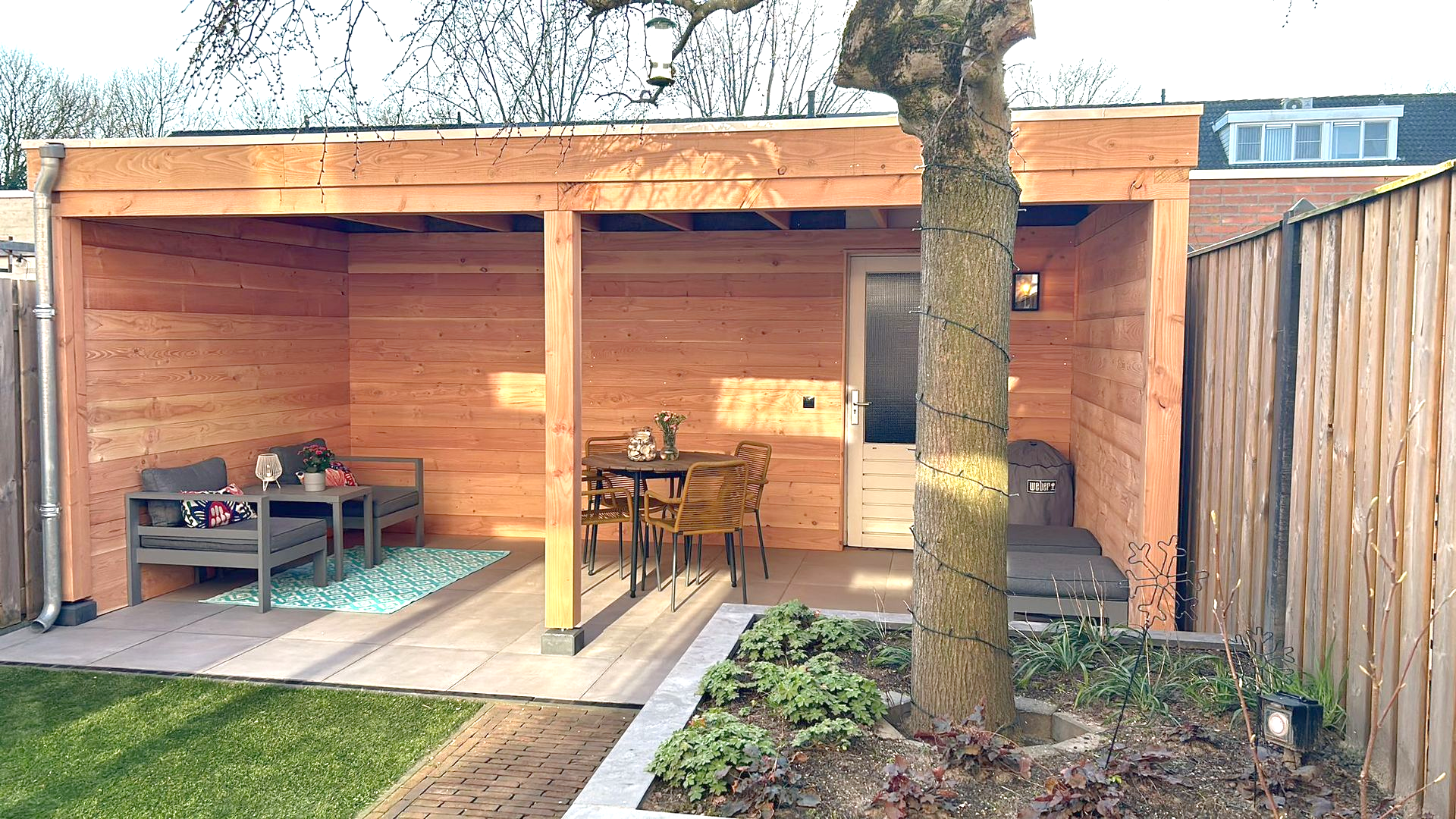 Houten buitenruimte met zitgelegenheid, tafel en deur. Boom op de voorgrond, omheinde tuin.