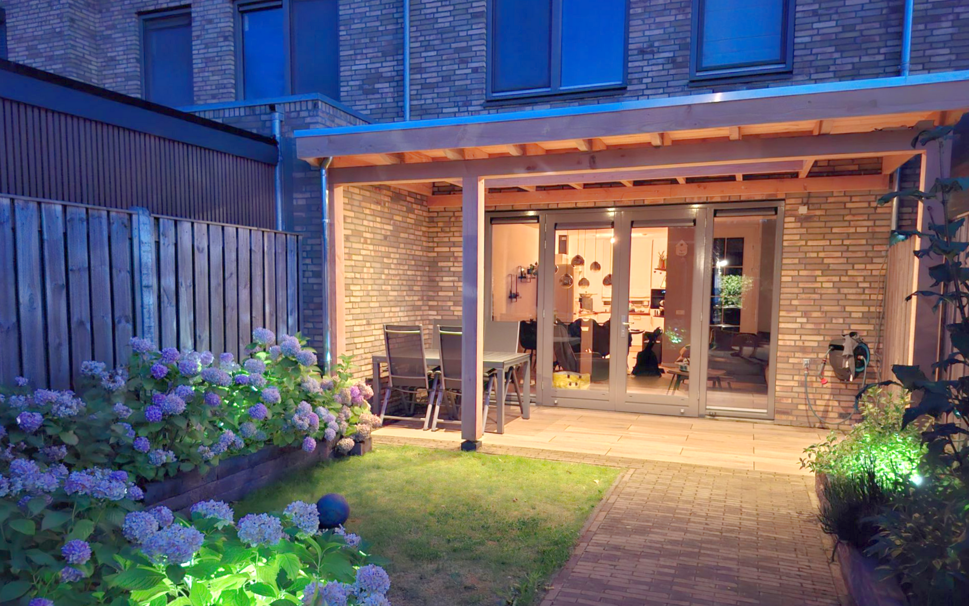 Terras in de achtertuin met houten balken. Schuifdeuren van glas leiden naar een interieur met verlicht gras en bloemen.
