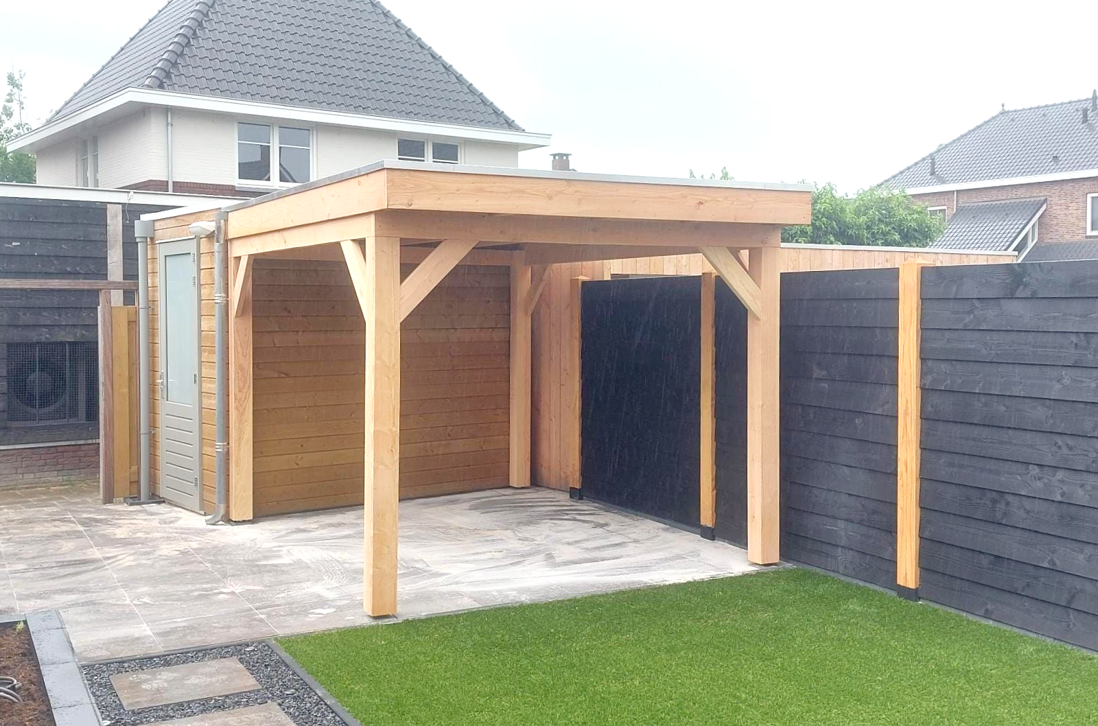 Houten carport met plat dak, betonnen vloer en aangrenzende zwarte schutting, in een achtertuin.