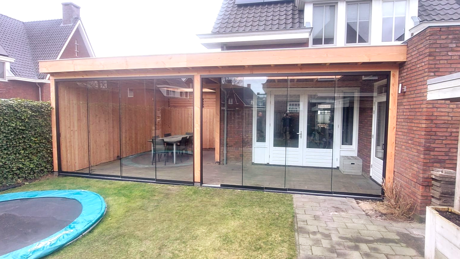 Houten terras met glazen wanden, grenzend aan een bakstenen huis, met een tafel, stoelen en een trampoline in de tuin.
