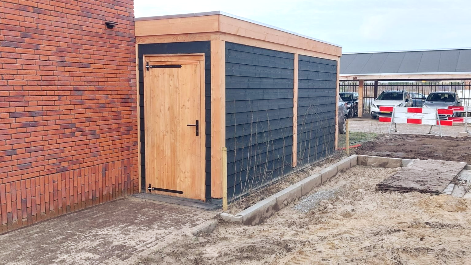 Houten schuur met zwarte gevelbekleding en een houten deur naast een bakstenen muur. Bouwplaatsomgeving.