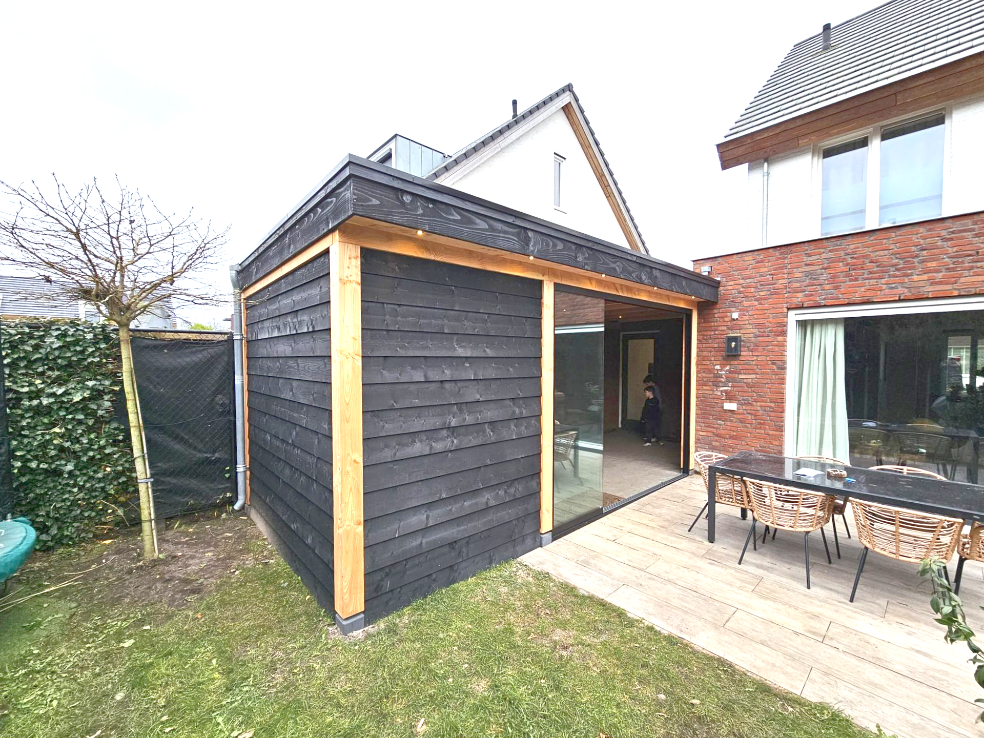 Zwart beklede tuinschuur met glazen schuifdeur, naast een bakstenen huis met een eethoek in de tuin.