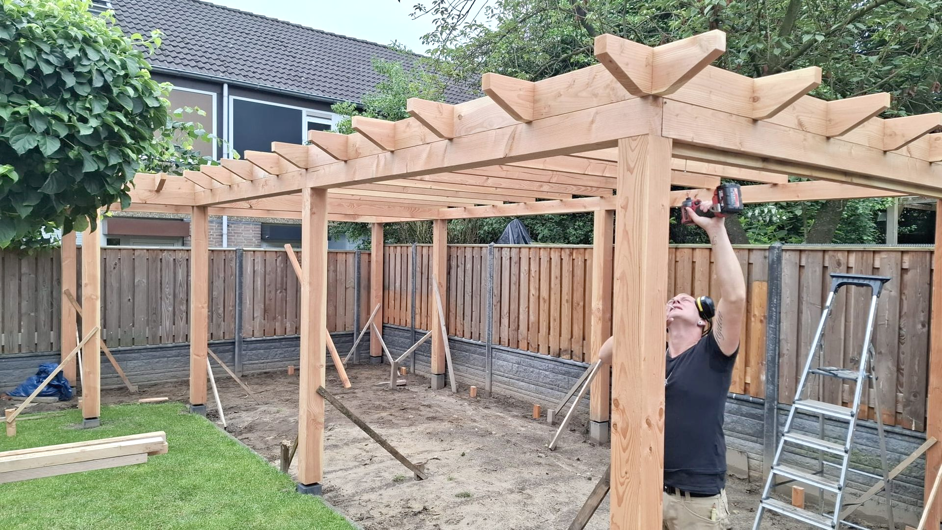 Een persoon die met behulp van een ladder een houten pergola in een achtertuin in elkaar zet.