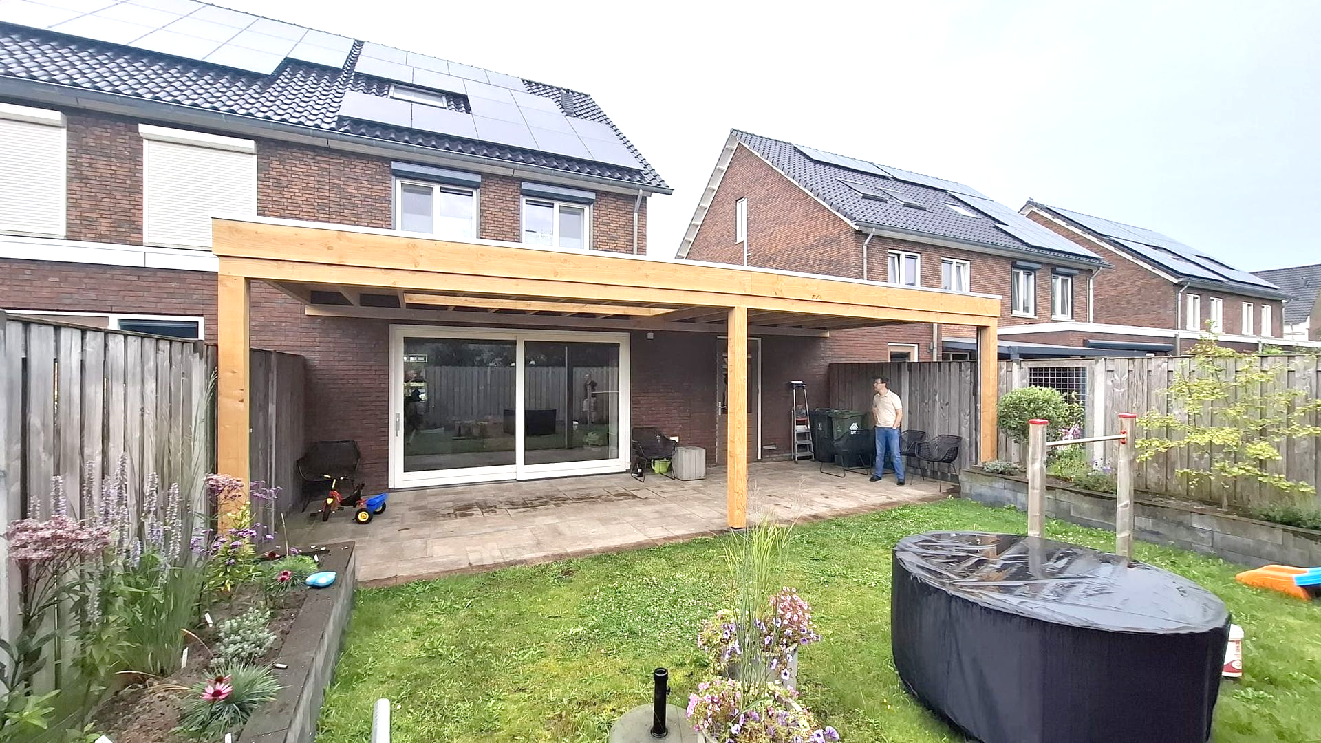 Achtertuin met houten pergola boven terras, persoon staat. Huizen op de achtergrond, groen gazon, bloemen en tuindecoratie.