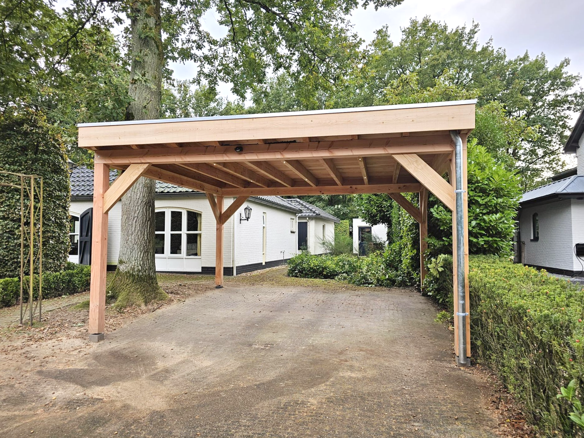 Carport met houten frame, oprit en witte gebouwen op de achtergrond.