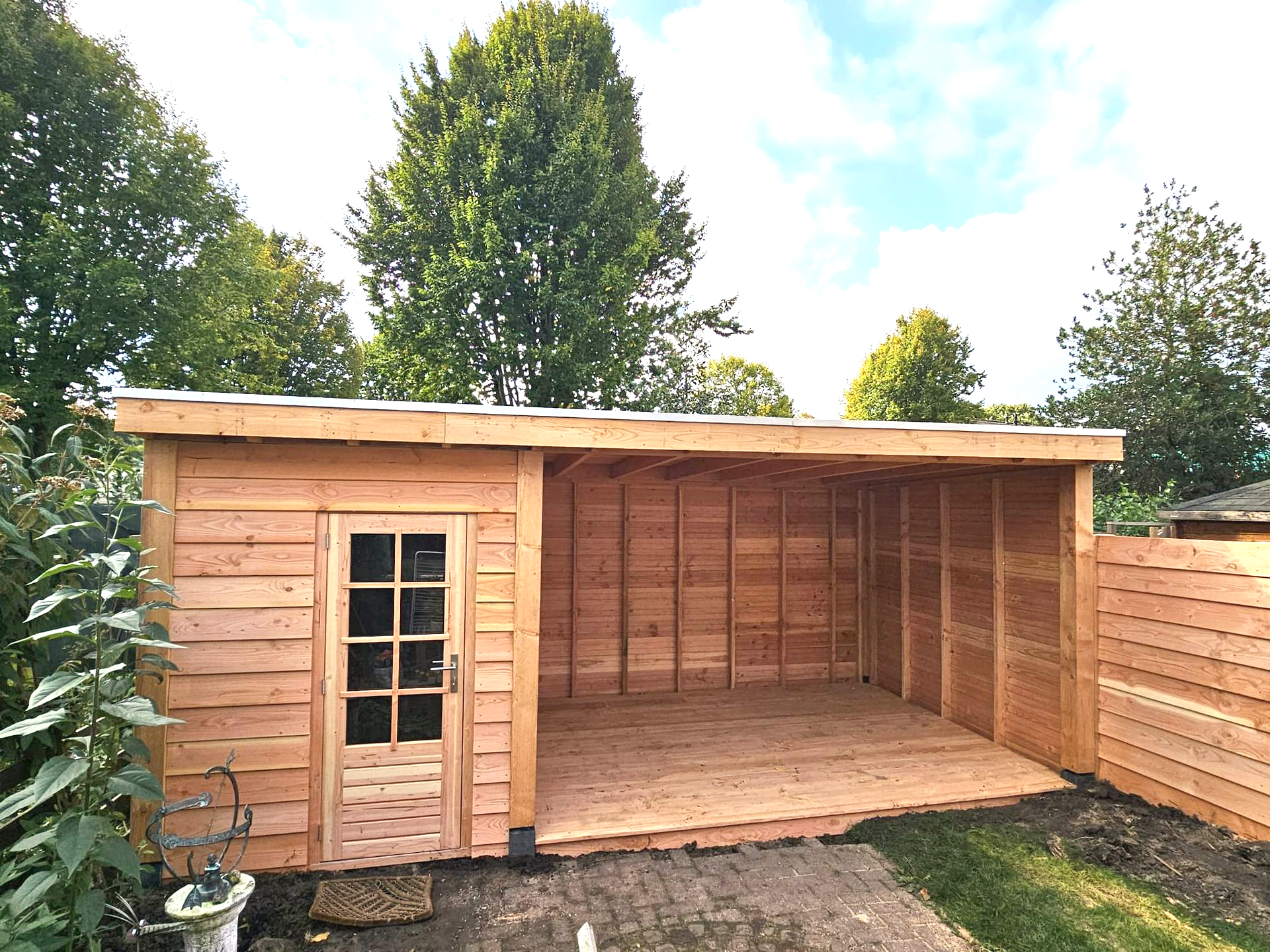 Houten schuur met een openluchtgedeelte; lichtbruine buitenkant. Gebouwd tegen bomen en een bewolkte hemel.