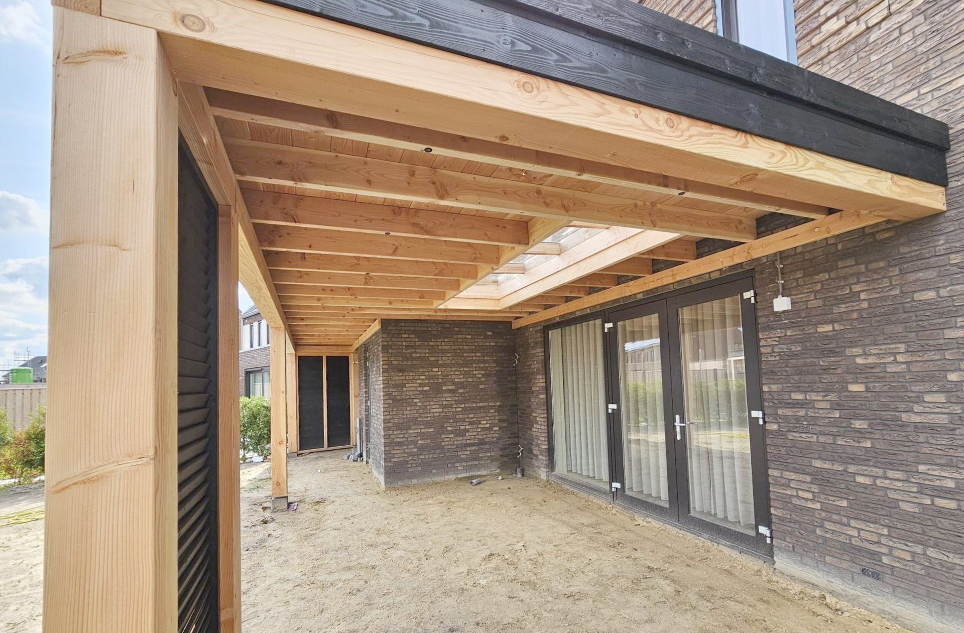 Overdekt terras met houten balken en een gedeeltelijk dakraam naast een bakstenen gebouw met glazen deuren.
