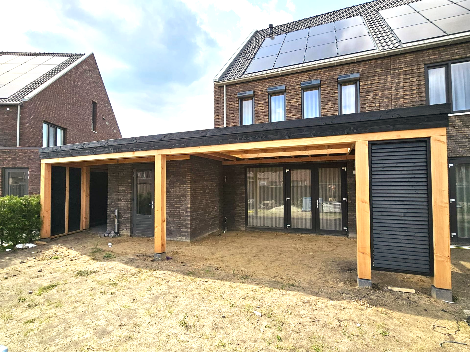 Een houten terrasoverkapping aan een bakstenen huis. Inclusief zonnepanelen, glazen deuren en een tuin van aarde.