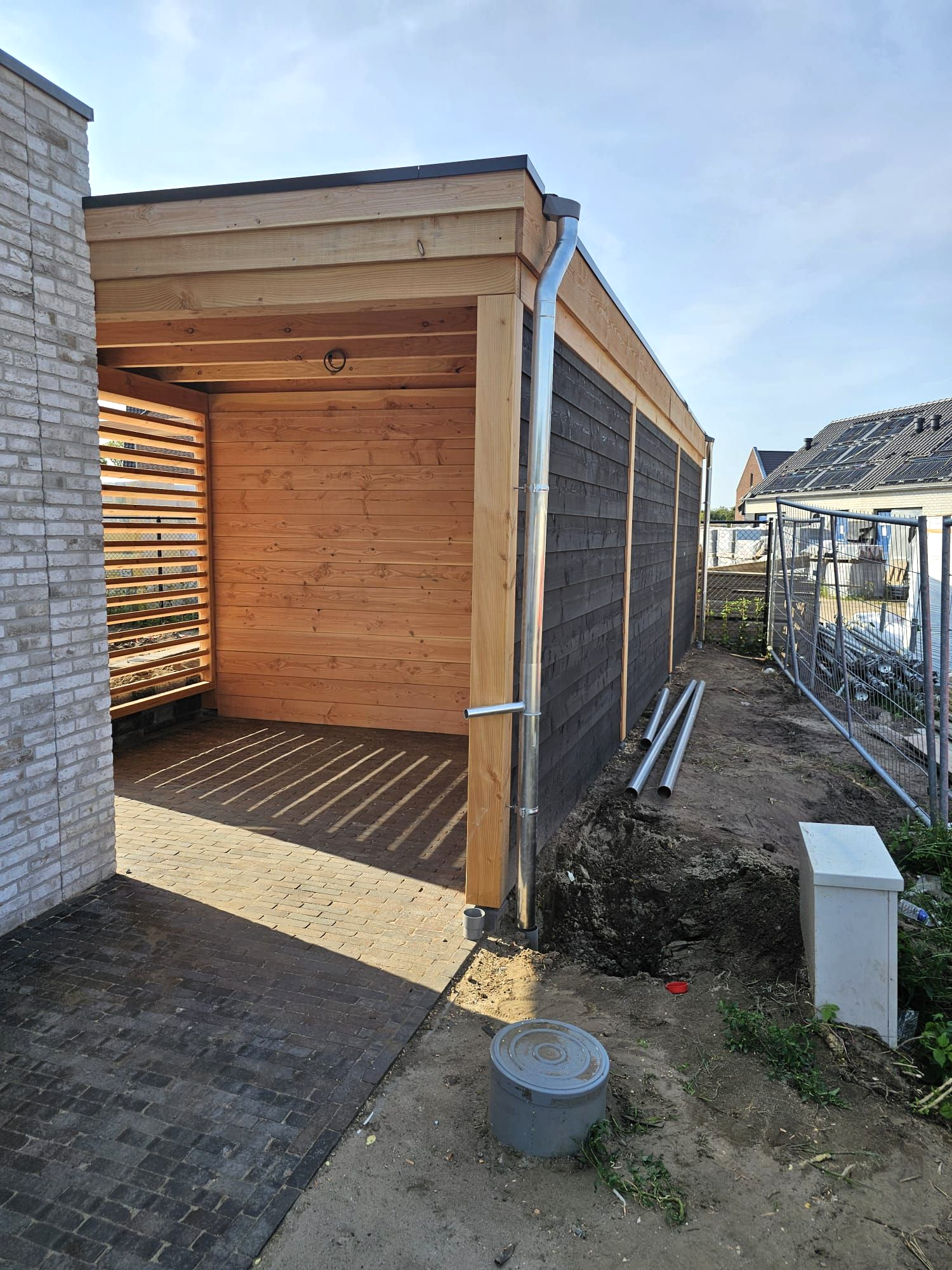 Houten carport met zwarte gevelbekleding, lichtgekleurd dak, dakgoot en betonnen pad.