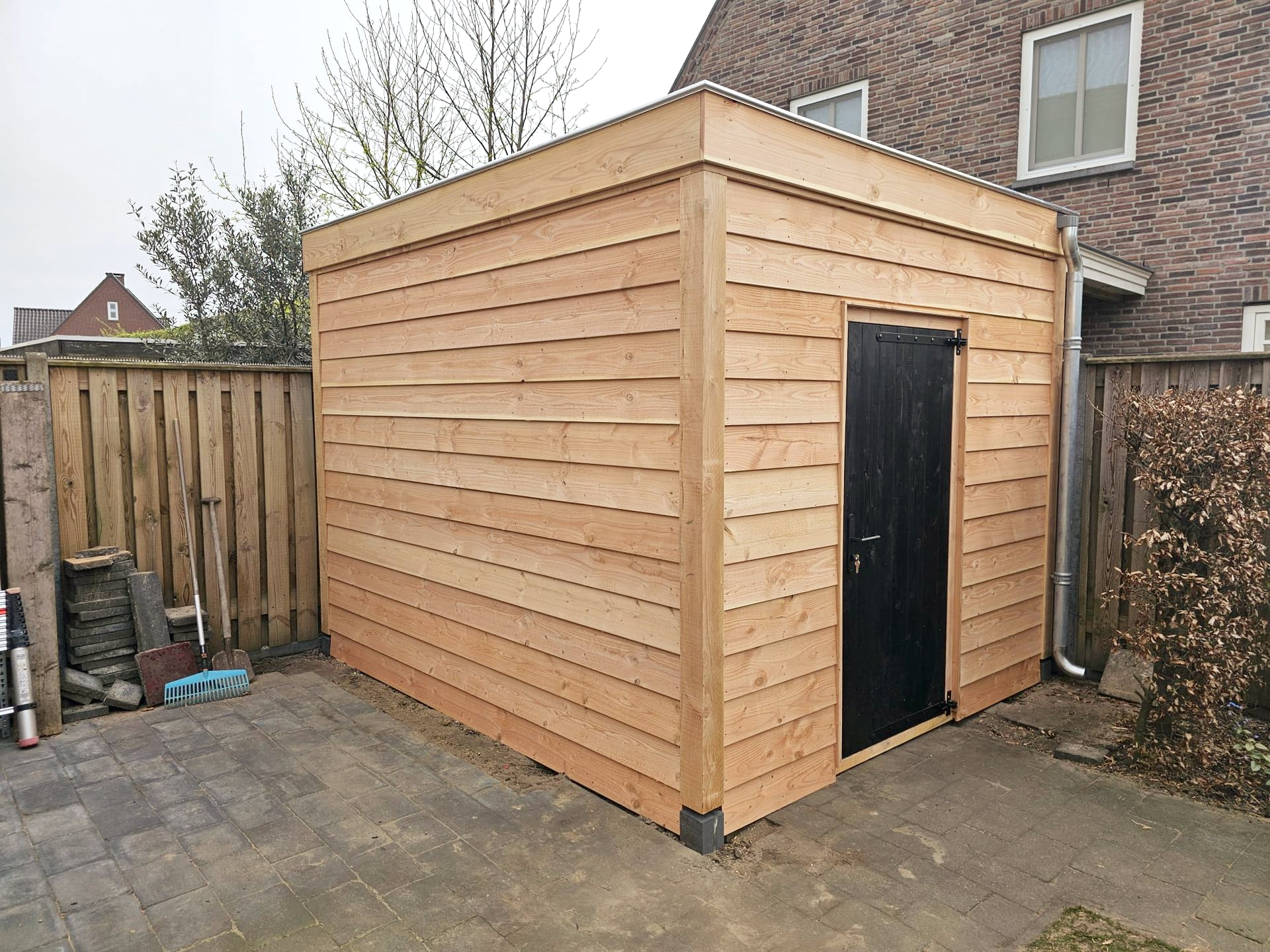 Houten schuur met zwarte deur in een achtertuin, omgeven door een hek en een bakstenen huis.