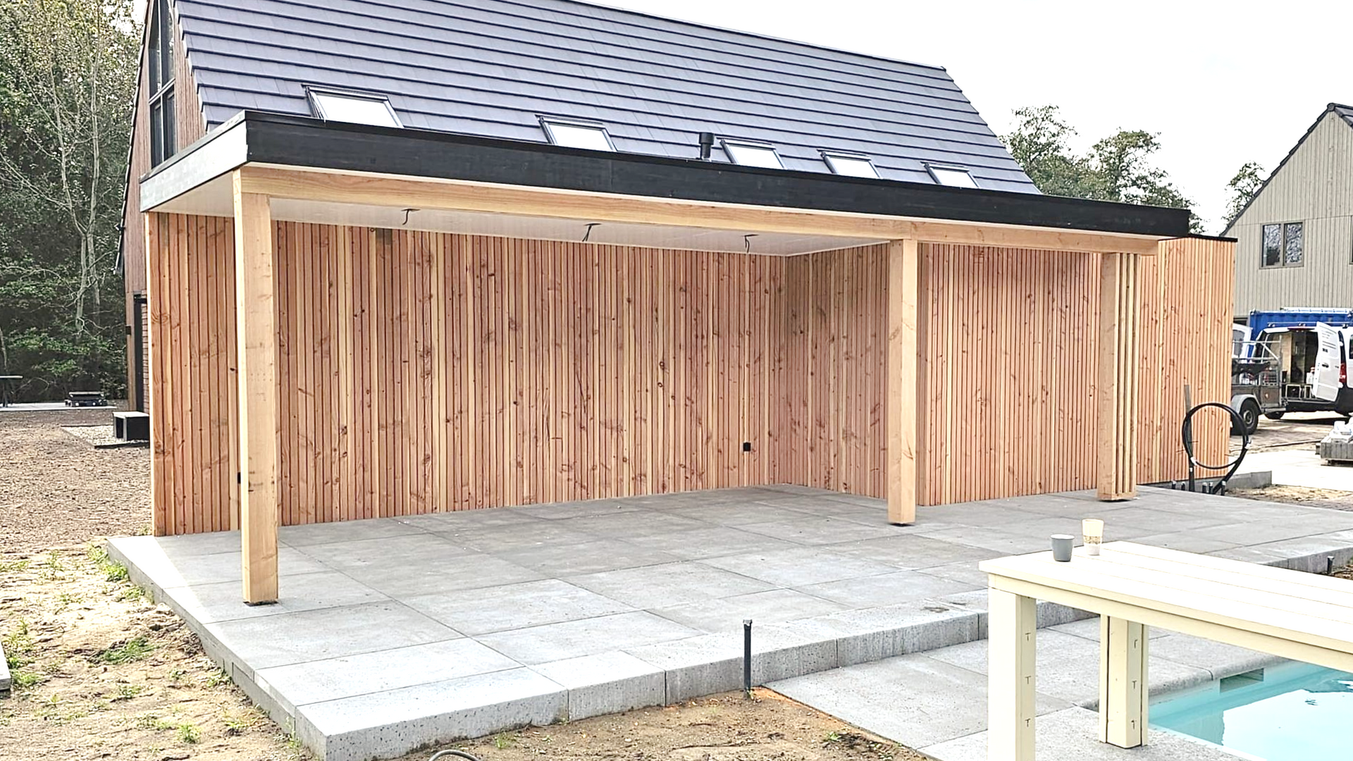 Houten terras met lichtgekleurde gevelbekleding, onder een constructie met een zwart dak, betonnen terras, vlakbij een huis.